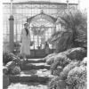 Image: Two women in pale skirts and jackets over dark blouses stand apart from each other outside a large glass conservatory building in this posed black and white photograph from the 1930s.