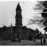 Image: A large, two-storey stone building with a tall clock tower protruding from one corner. A street full of people and automobiles is visible in the foreground