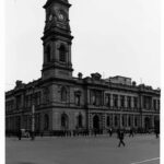 Image: A large, two-storey stone building with a tall clock tower protruding from one corner. A street and sidewalk with people standing on it is visible in the foreground