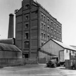 Image: A large, six-storey brick building with several windows on each floor and a large ventilation intake on its roof. Corrugated metal sheds stand in front and to the right of the building