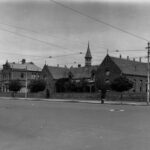 Image: a walled symmetrical stone building with a verandah with stone arches, a central spire, and two gable roofed wings with rose windows on the facade and double arched windows along the sides.