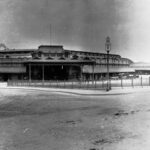 Image: the rear view of a railway station as seen from across a dirt road. The building features a corner entrance and a wrap around verandah.