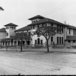 Image: A Spanish Mission-style two-storey building fronted by a dirt road and a row of small trees