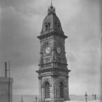 Image: A tall stone clock tower with an ornate roof and flagpole at its peak