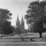 Image: A man in a suit and hat reclines on the grass in a park. A large stone church with twin spires is a prominent background landmark