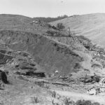 Image: A stone quarry cut into the side of a rolling hill. A wooden derrick is positioned adjacent to the top of the quarry