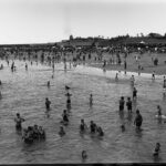 Image: View of beach with people bathing in the water and crowds on sand in the background