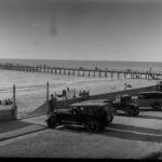 Image: View of jetty and beach with automobiles in the foreground in a parking lot.