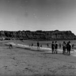 Image: Beach scene with people playing on the sand and in the water with rugged cliffs in the background