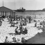 Image: People sunbathing on a beach with a jetty in the background.