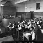 Image: Girls in black pinafores over white blouses sit at wooden desks with open books facing a blackboard. Under the blackboard, behind a large desk on a raised platform sits a woman, their teacher, also with an open book.