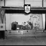 Image: Five teenagers, three boys and two girls, act out a scene on a small stage. The boys wear workers costumes, one girl is draped in a shawl and the other wears a full layered skirt, white blouse and vest. The props include a cook pot and gypsy wagon.