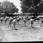 Image: A group of girls perform exercises in a grassy ground surrounded by trees and playground equipment