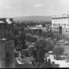 Image: A large six storey concrete building, with two storey columns flanking its entrance way, dominates one side of a tree lined street upon which a tram travels and 1930s era cars are parked.