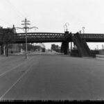 Image: two flights of stairs lead up to a metal bridge which passes over a wide paved road upon which an electric tram travels