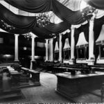 Image: wooden desks and leather bench seats are arranged around a room which is lined with white columns and decorated with portraits and swathes of dark fabric.