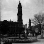 Image: A large, two-storey stone building with a tall clock tower protruding from one corner. A tree-lined park is visible in the foreground