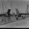 Image: A drawbridge within an active port is open to allow a tug to pass beneath it. A wharf with two mid-twentieth century sailing vessels tied alongside it is visible in the immediate foreground