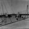 Image: A drawbridge within an active port is closed after a small boat has passed beneath it. A wharf with two mid-twentieth century sailing vessels tied alongside it is visible in the immediate foreground