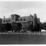 Image: A large, two-storey stone building with a central, square tower and steep, peaked roof