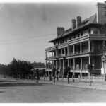 Image: A large, four-storey brick building sits alongside a dirt road lined with trees. Four horse-drawn carts are visible travelling along the street