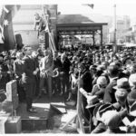 Image: a large crowd gathers around a man in a 1920s era dark suit who stands on am small stage with a carved foundation stone.