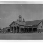 Image: a single storey stone building with pitched cross gable roof, protruding arched portico with verandahs on either side, and an open, octagonal  sided, domed cupola.