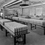 Image: A group of men and women stand in a large room full of rows of tables. Atop each table are several rows of artillery shells