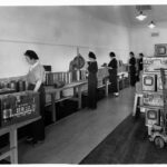 Image: A group of women stand at a long table manufacture and pack large-calibre artillery shells