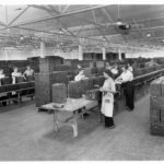 Image: A group of women and two men stand at tables in a large room and manufacture rectangular metal containers