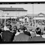 Image: A large crowd of people are gathered together at the Royal Adelaide Show, watching a wood chopping competition between 5 competitors on an open, outdoor space