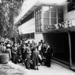 Image: A large group of British migrants of various ages arrive at hostel entrance. A sign above the door reads SPF Hostel.