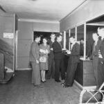 Image: six men and a woman in 1940s era dress stand around a reception desk while a seventh man leans on the desk, writing, behind a glass barrier. In the foreground a second woman in a hat sits looking at the group.