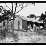 Image: A small group of young men and women sit on the porch of an old stone cottage. The cottage is surrounded by trees and scrub