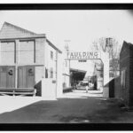 Image: A man in a white lab coat walks amongst a cluster of brick buildings. An archway in the foreground reads ‘Faulding Laboratories’