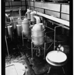 Image: A young Caucasian man in a white laboratory coat uses an industrial mixer to combine chemicals in a large steel vat. Large metal storage tanks are visible in the immediate background