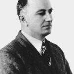 Image: A photographic head-and-shoulders portrait of a man wearing a suit with a striped tie