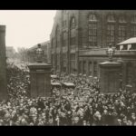 Image: A mass of men and women in 1920s attire walk through the entrance gates of a large, multi-storey brick factory