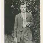 Bespectacled man in two piece suit and tie holding a pipe, and standing in a garden