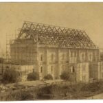 Image: A large hall is under construction. The stone walls are covered in scaffolding and the hammerbeams of the roof are visible. Men can be seen on the roof of the building.