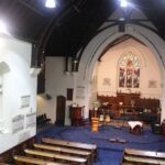 Image: An large, interior room with a high ceiling, wooden pews and a large stained glass window