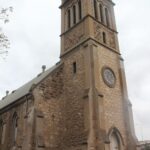 Image: the front entrance of a stone church with a single square tower with a clock.