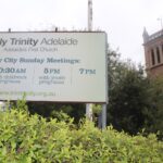 Image: A sign for Holy Trinity Church stands in front of the large stone church tower