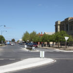 A large wide street in foreground extends into the distance. There is a large white stone building on the right hand corner