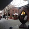 Image: steel quadrangle-shaped sculpture with rounded edges and a geometric hole in the middle in front of a brick courtyard with restaurants' patios