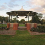 Image: A green and white gazebo with red brick footers stands in a park. A street with cars is visible in the left background