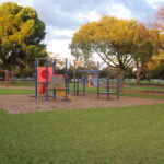 Image: Playground equipment sits in the middle of an open park interspersed with trees of different species