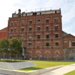 Image: A man and woman walk a dog in front of a tall, multi-storey historic brick building