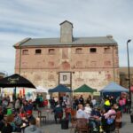 Image: A large group of people sit at tables and examine market stalls in front of a large, multi-storey historic stone building