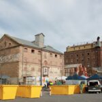 Image: A large group of people visit market stalls in front of two large, multi-storey historic buildings